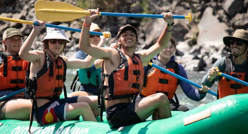 a group of students sitting in a raft smile and raise paddles in the air on an outward bound expedition